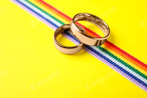 Preview: Wedding rings and rainbow ribbon on color background. Gay symbol