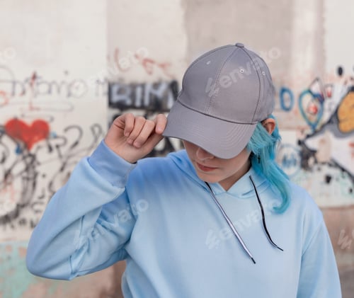 Preview: Blue haired Teenage girl in blue hoodie and baseball cap staying against graffiti wall