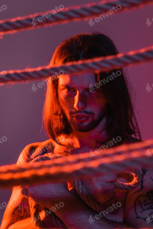 Preview: Bearded and long haired man looking at camera near blurred ropes on purple background with light