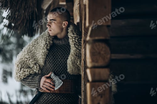 Preview: Ancient viking warrior with steel axe posing near wooden hut in scandinavian forest