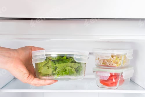 Preview: Female hand holds a glass box with green salad on the background of the freezer close-up.