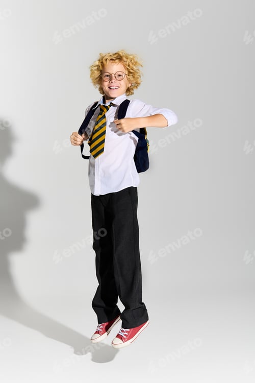 Preview: Blonde boy in school uniform joyfully jumping in a bright studio setting
