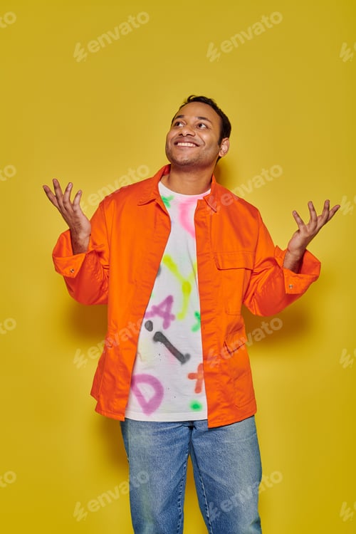 Preview: cheerful indian man in orange jacket and diy t-shirt gesturing on yellow backdrop, unbelievable