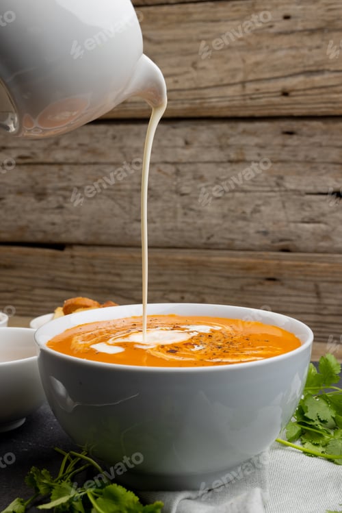 Preview: Vertical image of cream pouring into bowl of tomato soup, with wood background and copy space