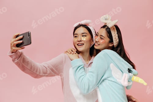 Preview: Tanned cool young women in pajamas take selfie on pink background. Happy girls with cosmetic eyepat