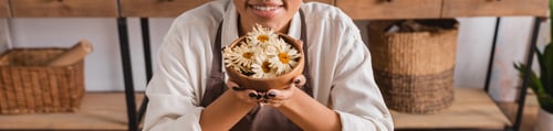 Preview: partial view of happy african american woman holding wooden bowl with dried camomiles in craft