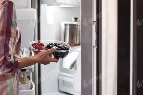 Preview: Woman is taking container with fresh berries from refrigerator