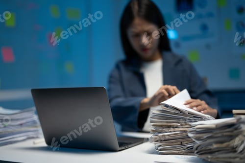 Preview: Overwhelmed businesswoman reviews paperwork late at night in office, exhausted and focused on loomin