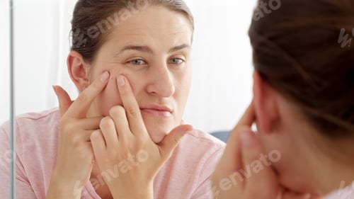 Preview: Young woman looking at her reflection and squeezing pimples and acne