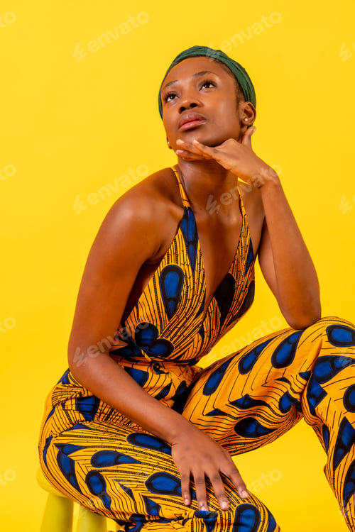 Preview: African young woman in traditional dress on yellow background sitting in studio looking at camera