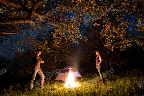 Preview: Tourists at a campfire near tent under trees and beautiful night sky full of stars. Night camping