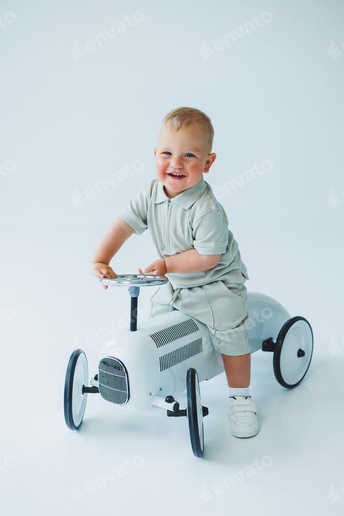 Preview: Stylish one-year-old boy in a suit with shorts and a T-shirt rides a car. Toddler boy on a white
