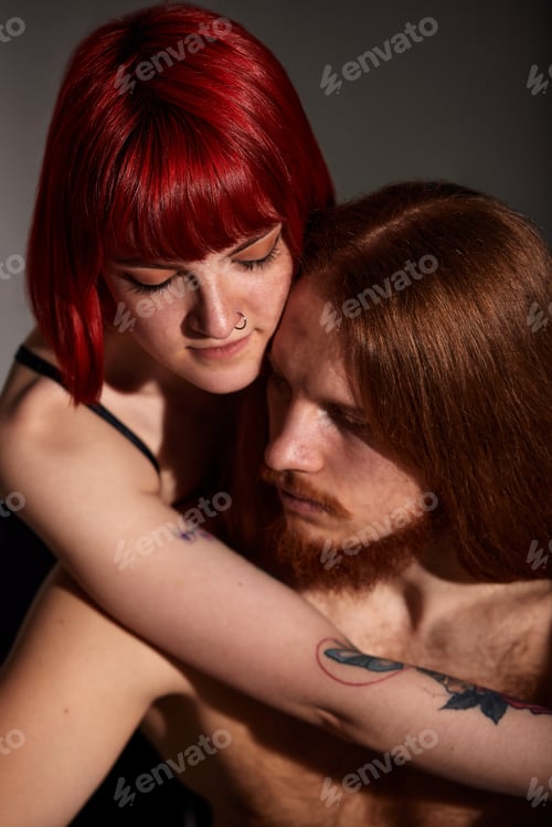 Preview: A young attractive woman with bright ruby hair, tattoos and piercings hugs her man with long hair