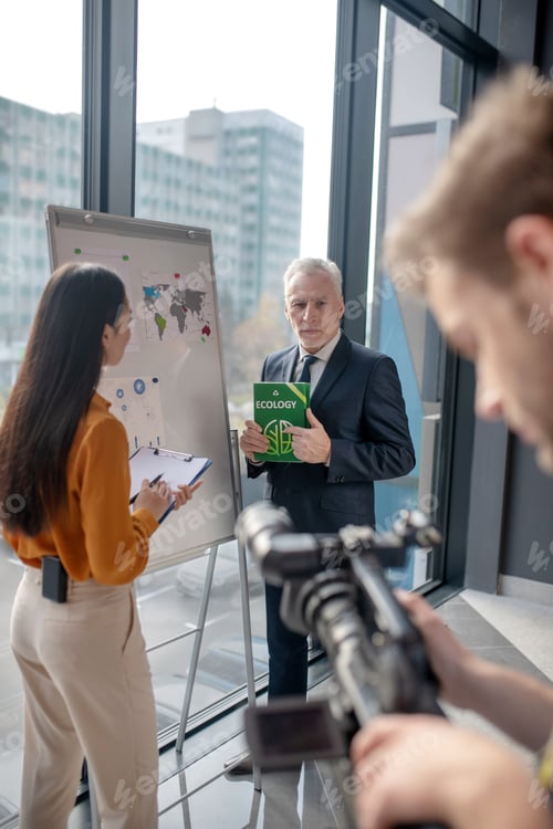 Preview: Grey-haired man talking to a young reporter in the studio