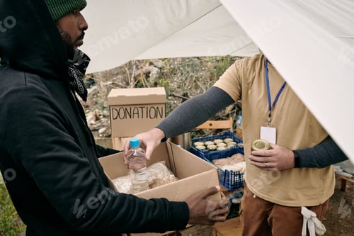 Preview: Young refugee man holding box while volunteer putting there food and water