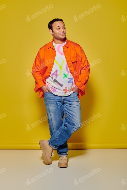 Preview: full length of stylish indian man in orange jacket and denim jacket posing on yellow background