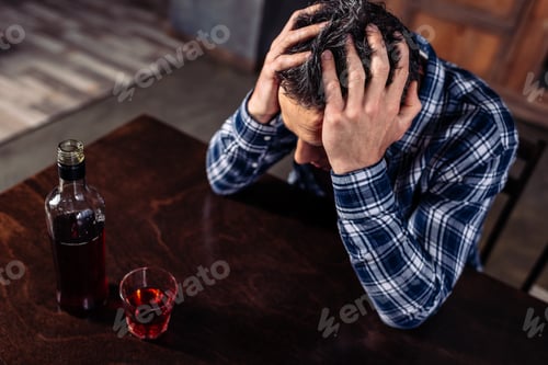 Preview: high angle view of drunk man sitting at table with bottle and glass of alcohol at home