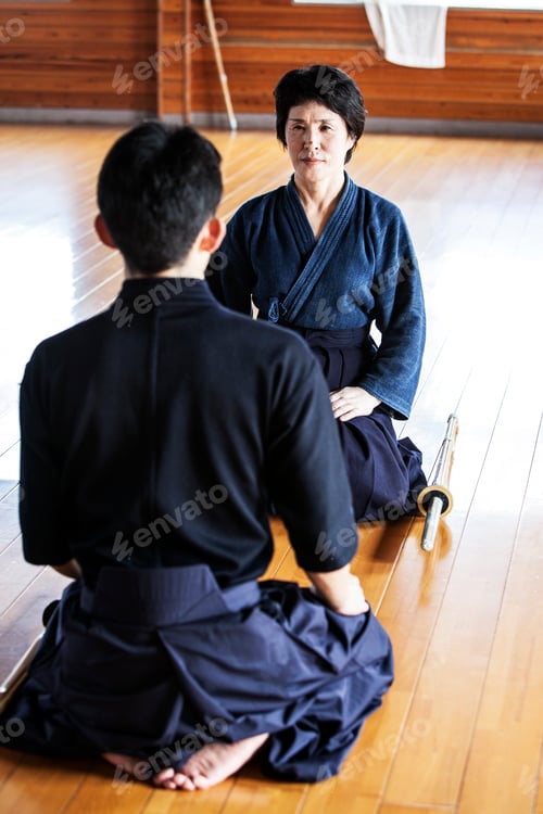 Preview: Female and male Japanese Kendo fighters kneeling opposite each other on wooden floor.