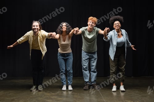 Preview: full length of happy multiethnic actors holding hands and bowing on stage of theater