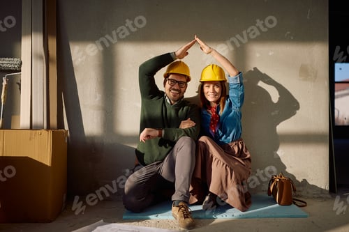 Preview: Happy couple making roof shape with their hands in new house and looking at camera.