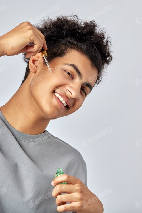 Preview: Young guy with black curly hair moisturizing his skin with a facial oil in an anti-aging skincare