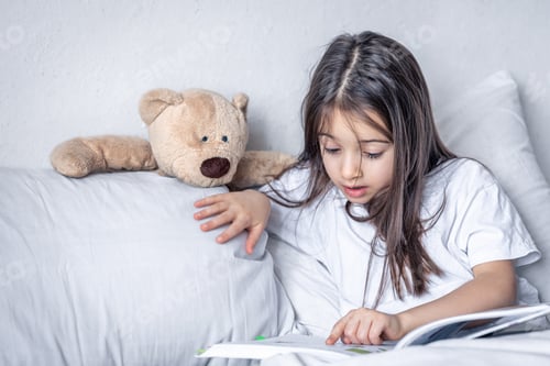 Preview: Little girl reads a book with a teddy bear in bed in the morning.