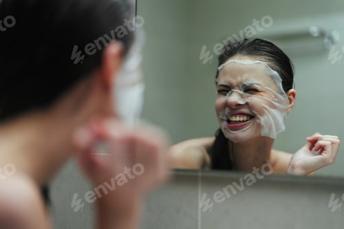 Preview: Joyful woman applying face mask in bathroom mirror, enjoying her skincare routine and self-care