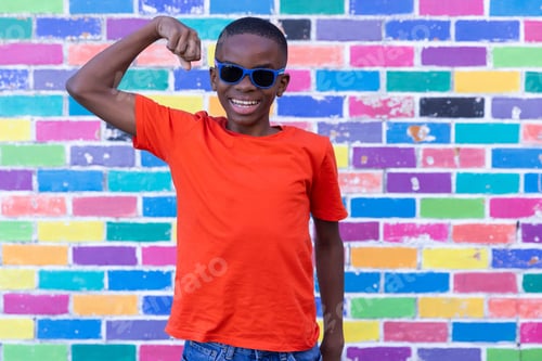 Preview: Smiling boy in sunglasses flexing arm muscles in front of colorful brick wall