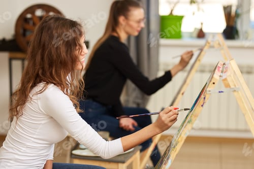 Preview: Girl with brown curly hair dressed in white blouse paints a picture at the easel in the drawing