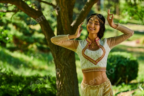 Preview: young and happy indian woman in authentic wear dancing in park on sunny day, summer happiness