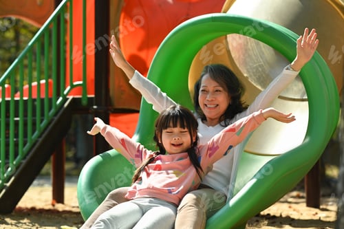 Preview: Cute little grandchild and mature grandma having fun on colorful slide.