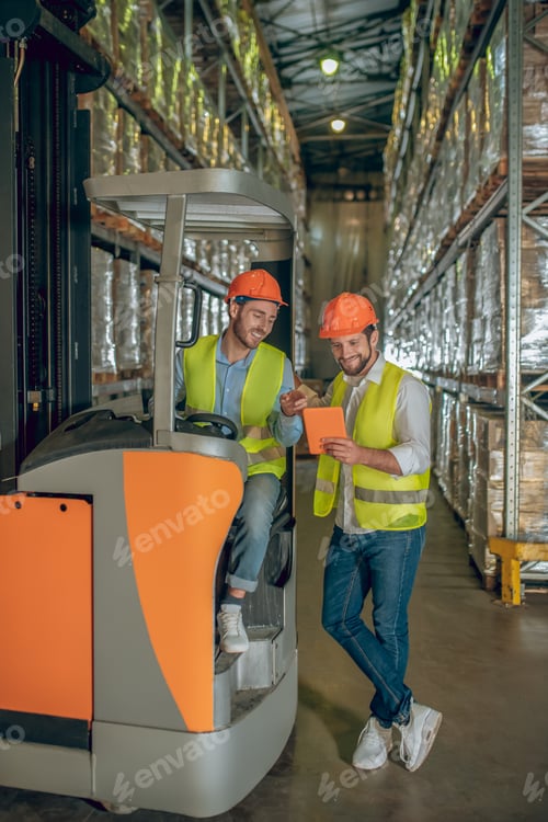 Preview: Conversation. Two Warehouse Workers In Helmets Looking At The Tablet And Discussing Something
