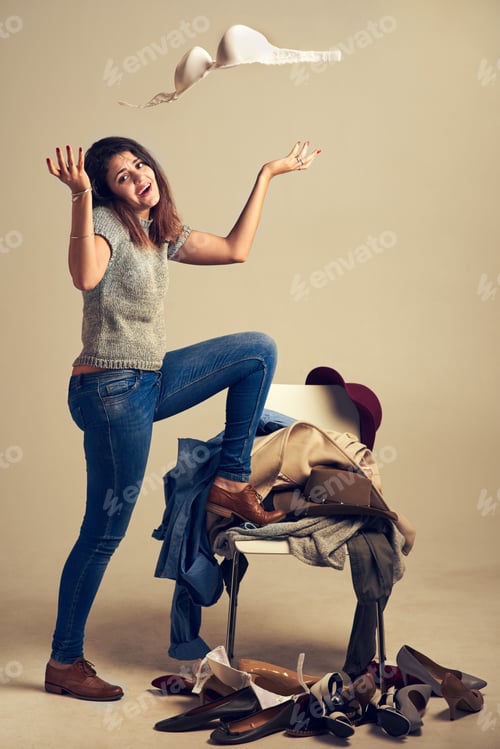 Preview: Studio shot of a young woman choosing clothing piled on a chair against a brown background