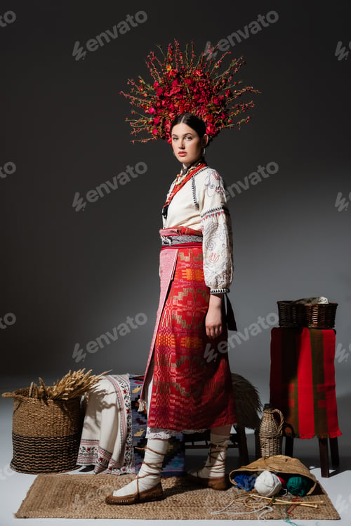 Preview: full length of ukrainian woman in traditional red floral wreath standing near yarn balls and wheat