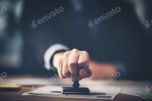 Vista previa: Close-up Of A Person's Hand Stamping With Approved Stamp On Document At Desk