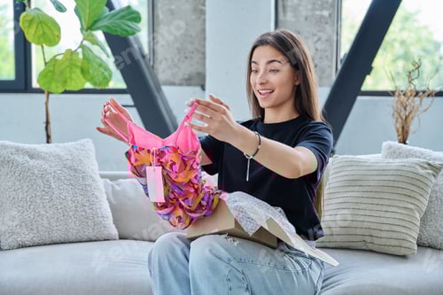 Preview: Young woman unpacking cardboard box parcel with online order, looking at new clothes