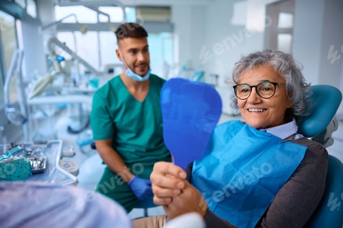 Preview: Happy senior woman looking her teeth in a mirror after dental procedure at dentist's office.