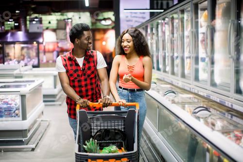 Preview: smiling african american couple with shopping trolley choosing food in supermarket