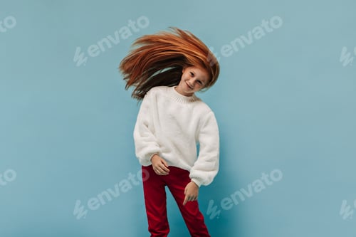 Preview: Wonderful girl in stylish white sweater and red pants smiling and playing her long ginger hair on i