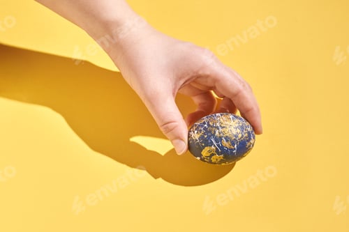 Preview: Caucasian adult hand holding blue and gold decorated easter egg on bright