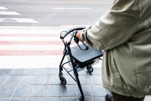 Vorschau: Ältester wartet darauf, einen Zebrastreifen zu überqueren, unterstützt von einem Spaziergänger.