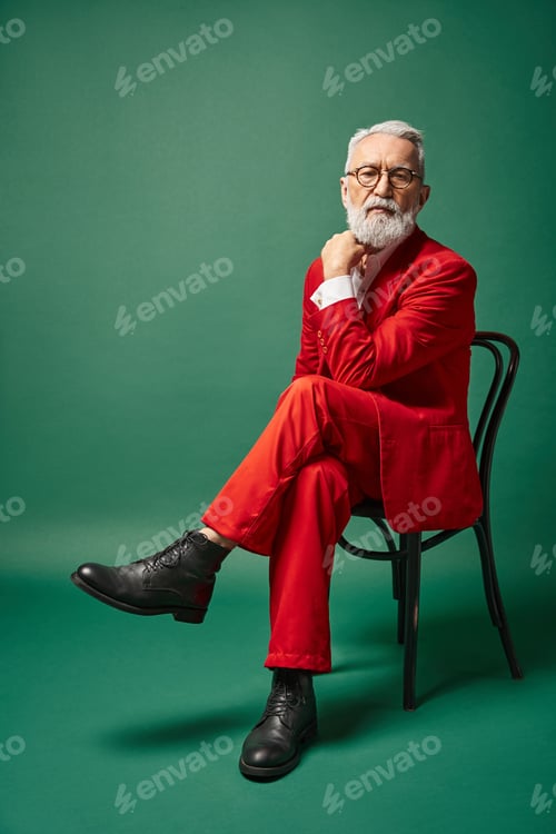 Preview: stylish man dressed as Santa with glasses sitting on chair with hand near face, winter concept