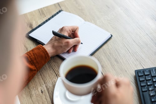 Preview: partial view of man writing in textbook and holding coffee cup at table with computer keyboard