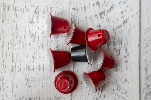 Preview: Vertical closeup shot of one black and many red coffee capsules on a weathered white wooden surface