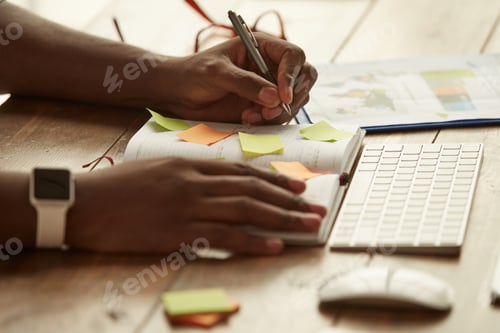 Preview: Cropped photo of afro american businessman making some notes while working remotely at home. Male