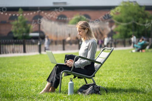 Preview: Relaxed focused woman with earbuds in grassy park using laptop. Streaming shows, music, movies