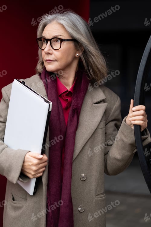 Preview: Elderly businesswoman with a folder stands confidently outside a modern office building during a