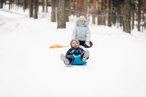 Preview: Happy boy sliding on snow in winter forest with sister watching him