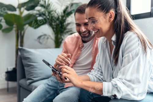 Vista previa: Feliz pareja joven mirando fotos en un teléfono inteligente mientras se divierte sentada en el sofá de casa.