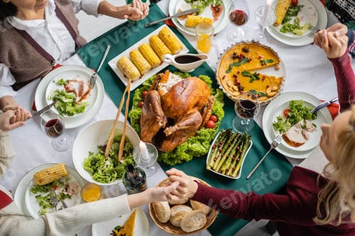 Preview: top view of interracial family holding hands while praying near tasty meal served for thanksgiving